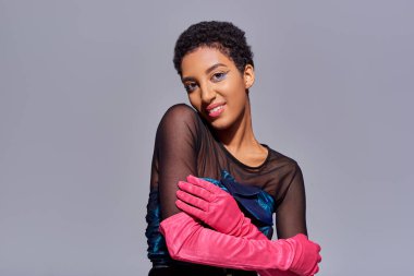 Gülümseyen ve modaya uygun Afro-Amerikan modeli kokteyl elbisesi ve pembe eldivenlerle koluna dokunuyor ve gri, modern nesil z moda konseptinde izole edilmiş bir şekilde kameraya bakıyor.