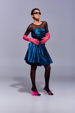 Neşeli ve şık Afro-Amerikan kadın güneş gözlüğü, kokteyl elbisesi ve topuklu ayakkabılar gri arka planda poz veren modern nesil z moda konsepti