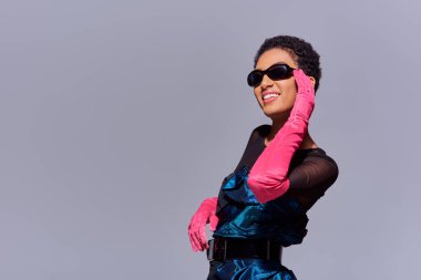 Joyful african american woman in cocktail dress and pink gloves touching sunglasses and standing isolated on grey, modern generation z fashion concept clipart