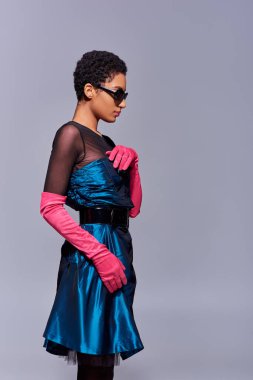 Kokteyl elbiseli, güneş gözlüklü ve pembe eldivenli modacı genç Afro-Amerikan kadın gri, modern nesil z moda konseptinde tek başına dikilirken yayına dokunuyor.