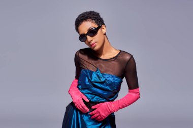 Güneş gözlüklü, kokteyl elbiseli ve pembe eldivenli özgüvenli ve zarif Afro-Amerikan modeli gri, modern nesil z moda kavramında izole edilmiş.