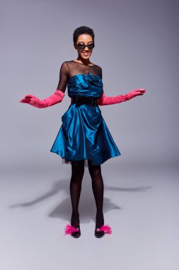 Full length of cheerful african american woman in sunglasses, cocktail dress and pink gloves posing in feathered shoes while standing on grey background, modern generation z fashion concept clipart