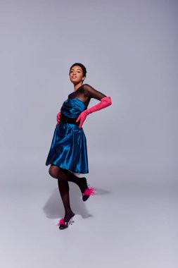 Trendy african american woman in cocktail dress, pink gloves and heels with feathers touching hips and smiling while standing on grey background, modern generation z fashion concept clipart