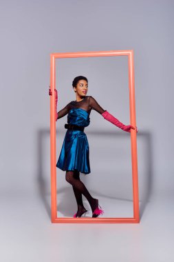 Trendy young african american model in cocktail dress, pink gloves and feathered shoes posing and standing near frame on grey background, modern generation z fashion concept clipart