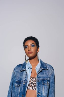 Portrait of confident african american model with bold makeup, denim jacket and top with animal print looking at camera while standing isolated on grey, denim fashion concept clipart