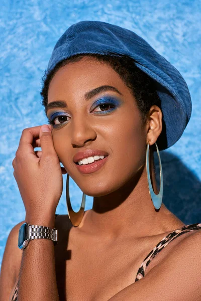 大胆なメイク ベレー帽 黄金のイヤリングを持つ若いアフリカ系アメリカ人女性の肖像画は カメラで笑顔と青のテクスチャの背景にポーズ スタイリッシュなデニムの衣装 — ストック写真