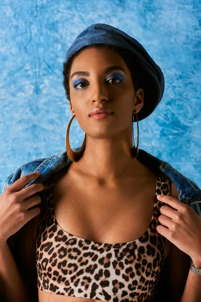 Confident young african american model in beret and golden earrings wearing denim jacket and top with leopard print on blue textured background, stylish denim attire
