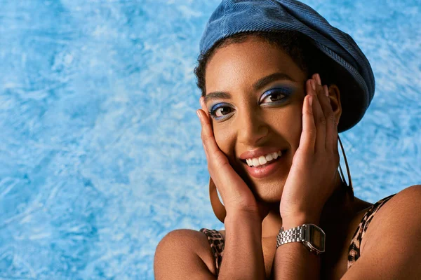 Portrait of trendy young african american model with bold makeup, earrings and beret touching cheeks and smiling at camera on blue textured background, stylish denim attire