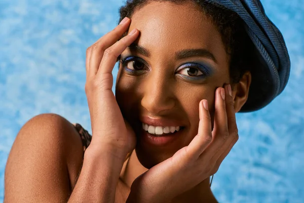 Close up view of smiling and young african american woman with vivid makeup and beret touching face and looking at camera on blue textured background, stylish denim attire