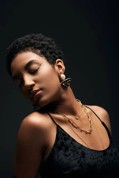 Fashionable african american woman with evening makeup, golden necklaces and dress closing eyes while standing isolated on black, high fashion and evening look