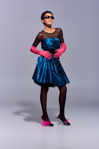 Full length of cheerful and stylish african american woman in sunglasses, cocktail dress and heels with feathers posing on grey background, modern generation z fashion concept