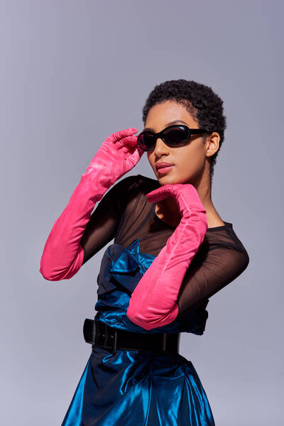 Portrait of elegant african american short haired woman in pink gloves, cocktail dress and sunglasses posing isolated on grey, modern generation z fashion concept