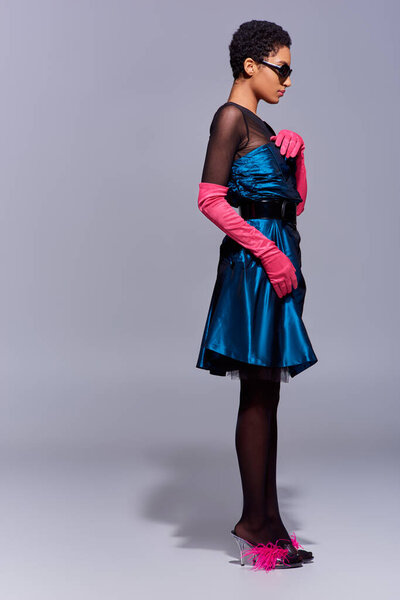 Full length of trendy african american woman in cocktail dress, pink gloves, feathered shoes and sunglasses standing on grey background, modern generation z fashion concept