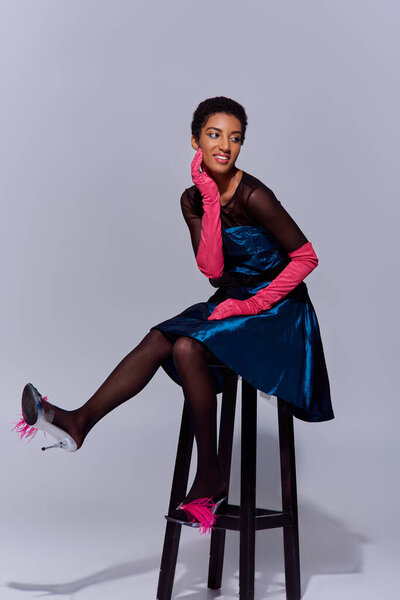 Joyful and stylish african american model in pink gloves, cocktail dress and heels with feathers posing while sitting on chair on grey background, modern generation z fashion concept