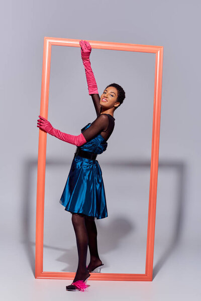 Full length of trendy african american woman in pink gloves, feathered shoes and cocktail dress smiling at camera while standing near frame on grey background, modern generation z fashion concept