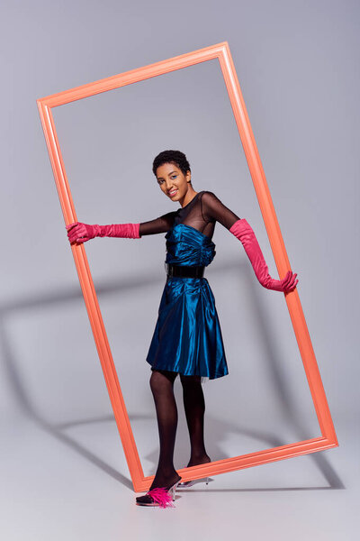 positive and confident african american model in cocktail dress, pink gloves and heels with feathers holding frame and standing on grey background, modern generation z, fashion concept