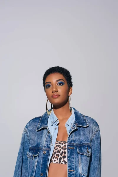 Portrait of confident african american model with bold makeup, denim jacket and top with animal print looking at camera while standing isolated on grey, denim fashion concept