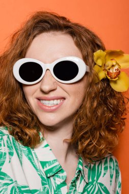 Orkide çiçekli, güneş gözlüğü ve bluzla poz veren, turuncu arka planda çiçek desenli genç bir kadının portresi. Yaz gündelik ve moda konsepti, gençlik kültürü.