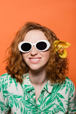 Smiling young woman with orchid flower in red hair wearing sunglasses and blouse with floral print while standing isolated on orange, stylish casual outfit and summer vibes concept, Youth Culture clipart