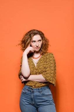 Pensive young red haired woman in modern yellow blouse and jeans looking away while standing and posing on orange background, stylish casual outfit and summer vibes concept, Youth Culture clipart