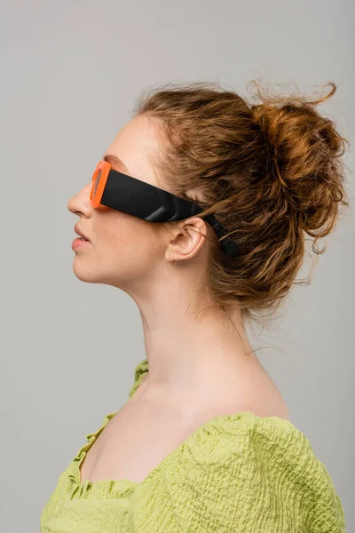 Yeşil bluzlu, güneş gözlüklü, doğal makyajlı modern kızıl saçlı kadın gri arka planda, modaya uygun güneş koruması konsepti, manken 