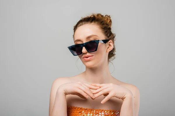 stock image Young red haired woman in top with orange sequins and naked shoulders posing in sunglasses and standing isolated on grey background, trendy sun protection concept, fashion model 