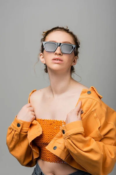 stock image Fashionable red haired model in sunglasses and orange top with sequins touching denim jacket while standing and posing isolated on grey background, trendy sun protection concept, fashion model 