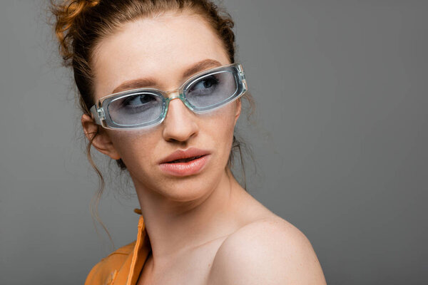 Модная молодая рыжеволосая женщина с естественным макияжем и причудами в синих солнцезащитных очках, которая отворачивается, стоя в изоляции на сером фоне, триумфальная концепция защиты от солнца 