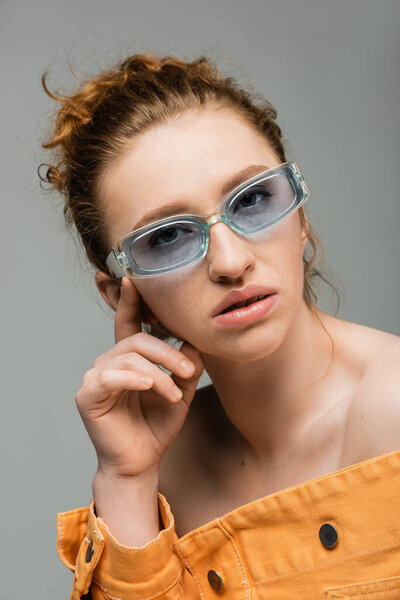 Портрет уверенной молодой рыжей женщины в солнцезащитных очках и оранжевой джинсовой куртке с обнаженными плечами, смотрящей на камеру на сером фоне, модная концепция защиты от солнца, модель 