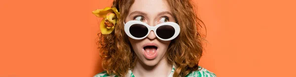 stock image Young and shocked redhead woman with orchid flower and stylish sunglasses looking away while standing on orange background, summer casual and fashion concept, banner, Youth Culture