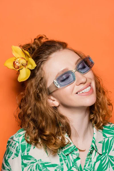 赤髪とランの花を持つ笑顔と流行の若い女性は サングラスやブラウスに花のプリントとオレンジの背景にポーズ 夏のカジュアルとファッションのコンセプト 青少年文化 — ストック写真