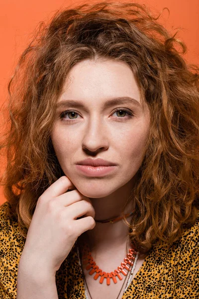 stock image Portrait of young redhead woman with natural makeup wearing necklaces and blouse with abstract print looking at camera isolated on orange, stylish casual outfit and summer vibes concept 