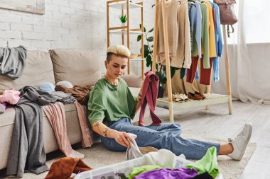 tattooed woman sorting wardrobe items while sitting on floor in living room near couch and rack with clothes, decluttering and reducing, sustainable living and mindful consumerism concept clipart
