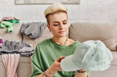 Gülümseyen bir kadın kanepenin yanındaki kıyafetleri seçerken şapkaya bakıyor, olumlu duygular, moda saç stili, dövme, sürdürülebilir yaşam ve dikkatli tüketim konsepti