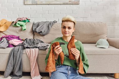 sorting garments, young woman with trendy hairstyle and tattoo holding suede boots, looking at camera near garments on couch in modern living room, sustainable living and mindful consumerism concept clipart