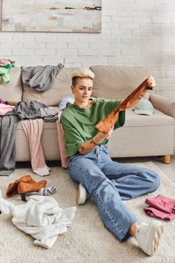 tattooed woman in casual style clothes holding suede boot while sorting belongings and thrift store finds near couch on floor in living room, sustainable living and mindful consumerism concept clipart