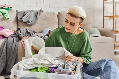Dövmeli ve modaya uygun saç stili olan genç bir kadın modern oturma odasındaki kanepenin yanındaki plastik konteynırda ikinci el kıyafetleri sınıflandırıyor, sürdürülebilir yaşam ve dikkatli tüketim konsepti