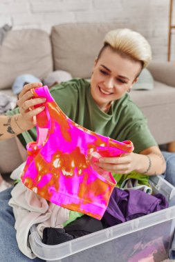 happy emotion, joyful woman sorting second-hand clothes and holding colorful top, trendy hairstyle, tattoo, blurred background, sustainable living and mindful consumerism concept clipart