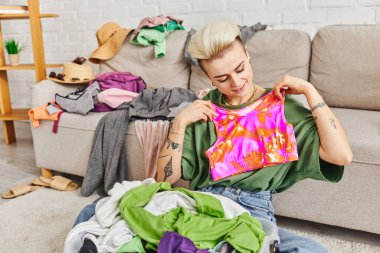 smiling woman holding colorful top while decluttering wardrobe items near couch with clothing in modern living room, trendy hairstyle, tattoo, sustainable living and mindful consumerism concept clipart