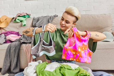 pleased woman looking at colorful tops near second-hand clothes and couch, reducing wardrobe, home decluttering, trendy hairstyle, tattoo, sustainable living and mindful consumerism concept  clipart