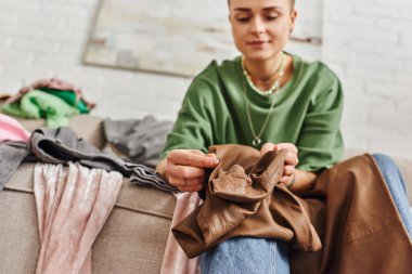 low angle view of young woman holding stylish leather pants while sitting on couch in living room and sorting clothes at home,  blurred background, sustainable living and mindful consumerism concept clipart