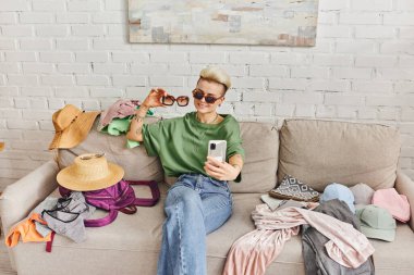positive tattooed woman in casual clothes and stylish sunglasses taking selfie on couch near pre-loved and wardrobe items for online exchange, sustainable living and mindful consumerism concept clipart