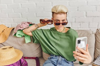 stylish tattooed woman with sunglasses taking selfie on smartphone near straw hats and clothing on couch, online swap, virtual marketplace, sustainable living and mindful consumerism concept clipart