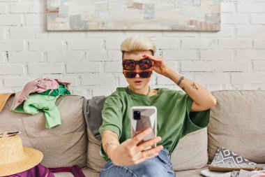 Genç ve stil sahibi güneş gözlüklü bir kadın akıllı telefonda hasır şapka ve giysi yanında selfie çekiyor. Evdeki kanepe, internet alışverişi, sürdürülebilir yaşam ve düşünceli tüketim kavramı.