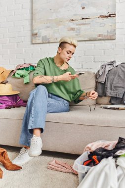 online swap on virtual marketplace, tattooed woman in casual clothes taking photo of sunglasses on couch near wardrobe items in modern living room, sustainable living and mindful consumerism concept clipart