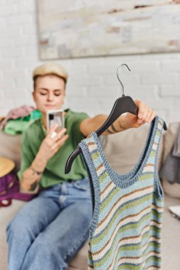 Bulanık tişörtlü genç bir kadın internet pazarında çevrimiçi takas için akıllı telefonla fotoğraf çekiyor. Giysileri düşüyor. Sürdürülebilir yaşam ve düşünceli tüketim kavramı.