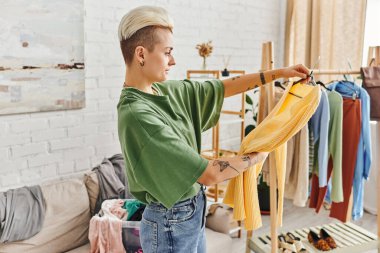 side view of tattooed woman looking at yellow jumper in living room, couch and rack with clothes, wardrobe items sorting and reducing, sustainable living and mindful consumerism concept clipart
