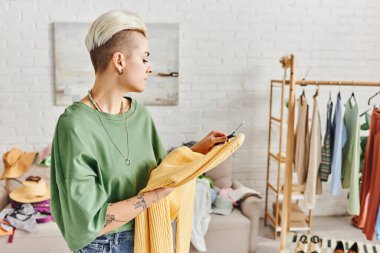 stylish tattooed woman looking at yellow jumper near rack with clothing and footwear in modern living room, wardrobe items sorting, sustainable living and mindful consumerism concept clipart