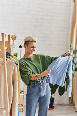 tattooed and satisfied woman looking at blue cardigan while sorting clothes near rack with wardrobe items on hangers in living room at home, sustainable fashion and mindful consumerism concept clipart