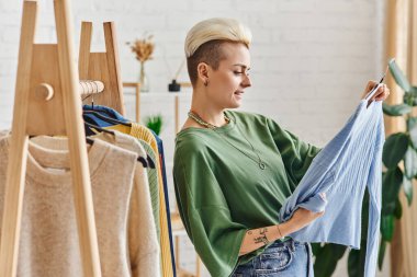 Giysi tasnif etme, moda ve dövmeli bir kadının askılarda gardırop eşyaları, sürdürülebilir moda ve dikkatli tüketim konsepti olan mavi hırkaya bakması.